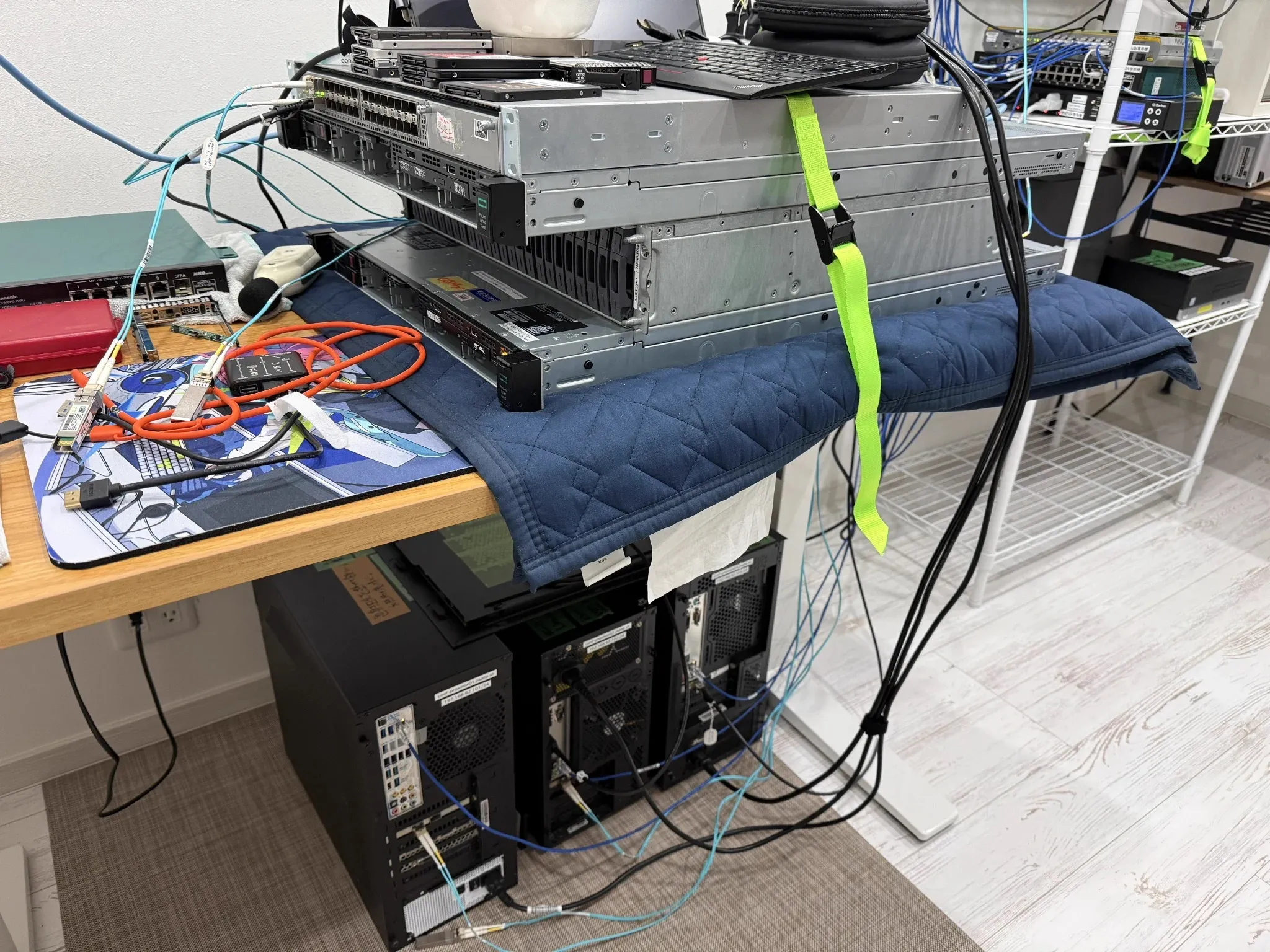 机の上に積み重ねたラックマウントサーバーとスイッチ。机の下にはタワー型のサーバーが並び、光ファイバーと LAN ケーブルが何本も伸びている