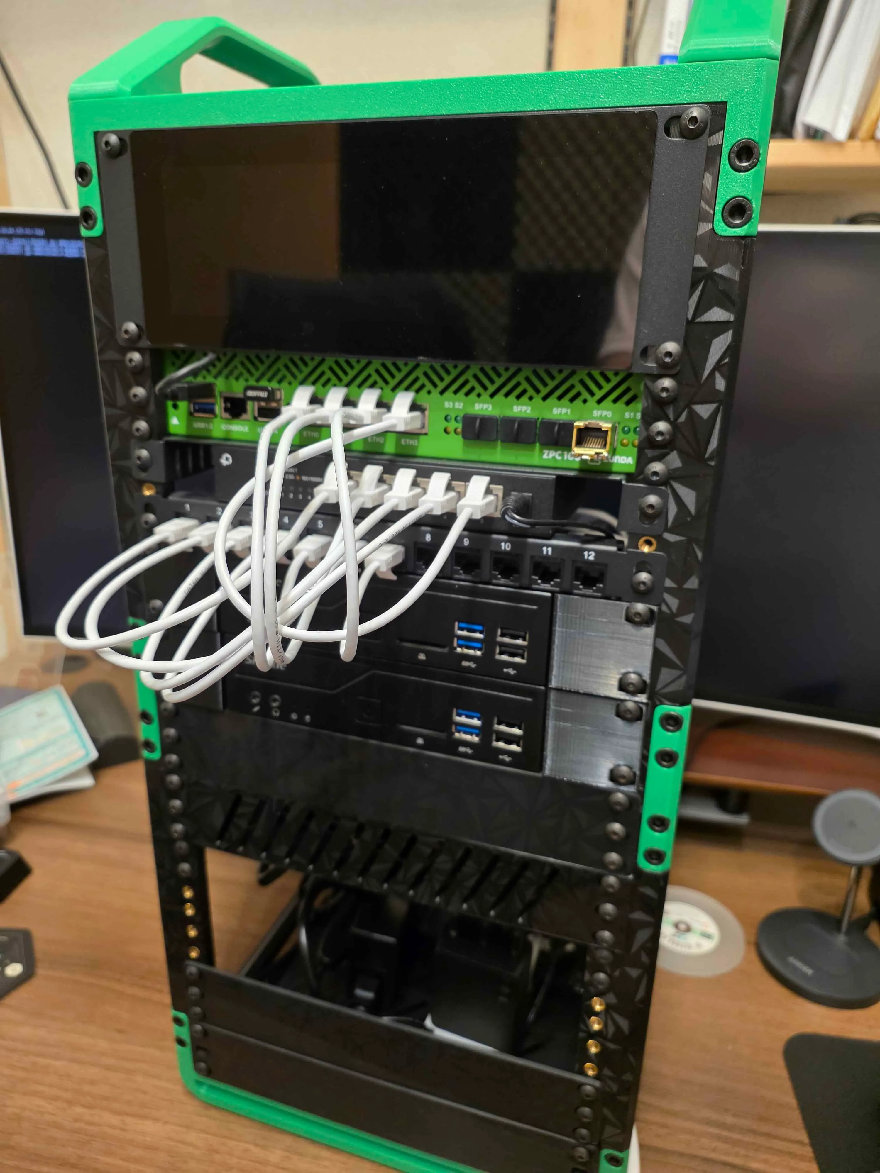緑と黒の小型ラック。上から液晶パネル、ルーター、スイッチ、パッチパネル、ミニ PC が収まり、白い LAN ケーブルでつながっている